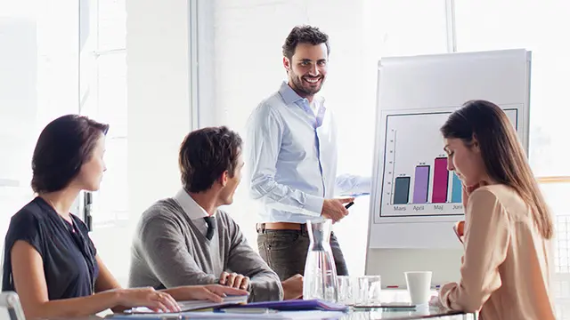 Personer sitter runt ett bord på ett möte och lyssnar på en som presenterar